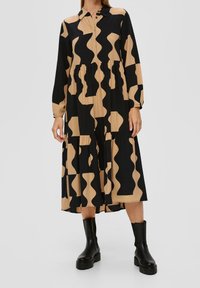 Abito a strati nero e beige con motivo ondulato, apertura con bottoni, maniche lunghe e colletto da camicia. Realizzato in tessuto leggero. Stivali neri indossati.