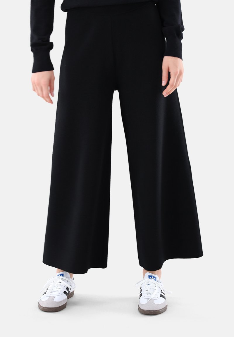 Schwarze weit geschnittene Hose aus weichem Stoff mit einem elastischen Bund. Kombiniert mit weißen Sportschuhen für einen lässigen Look.