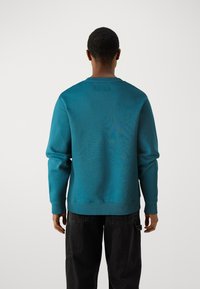 Tyrkis sweatshirt med ribbede mansjetter og kant, design med rund hals, glatt tekstur, og en merkelapp bak. Brukt med mørke bukser.