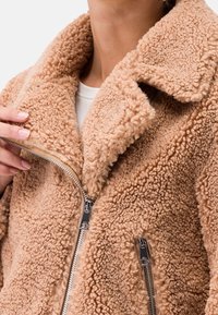 Persona che indossa una giacca in fleece sherpa beige con un ampio colletto, mentre tiene la zip vicino alla parte superiore, sopra una maglietta bianca.