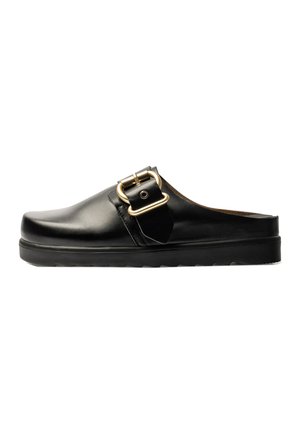 Zueco de cuero negro tipo slip-on con banda ancha y gran hebilla dorada en el lateral, suela plana con fondo texturizado.
