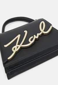 Schwarze Lederhandtasche mit einem auffälligen goldenen Schriftlogo. Das Design ist rechteckig mit einem oberen Griff und gesteppten Akzenten.