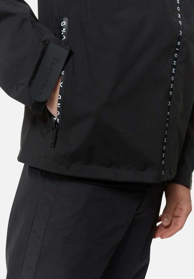 Giacca impermeabile nera con colletto alto, tasca con cerniera e dettagli del logo lungo la cerniera. Tessuto a trama liscia con design aderente.