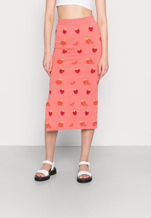 Femme portant une jupe rose mi-longue avec des motifs de fraises, d'oranges et d'agrumes, associée à des sandales blanches et un crop top blanc.