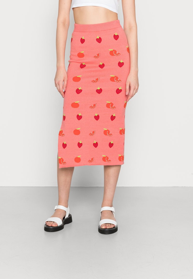 Femme portant une jupe rose mi-longue avec des motifs de fraises, d'oranges et d'agrumes, associée à des sandales blanches et un crop top blanc.