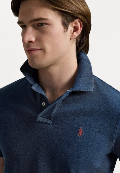 Navy zila polo krekls no teksturētas auduma, ar pogājamām apkaklēm un mazu sarkanu logo kreisajā krūtī.