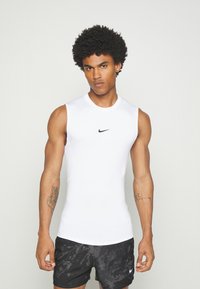 Bela brezrokavna atletska majica iz gladkega in elastičnega materiala, ki ima majhen črn Nike logotip na prsih. Nosimo jo s patterned črnimi kratkimi hlačami.