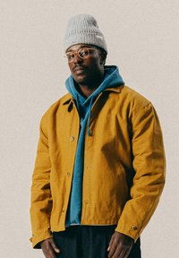 Άνδρας που φορά στρογγυλά γυαλιά, γκρι πλεκτό καπέλο, μπλε φούτερ και μπουφάν μουστάρδι κίτρινο, στέκεται μπροστά σε απλή φόντο.