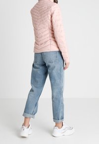 Jasnoróżowa pikowana kurtka z przeszyciami w kształcie litery V, zestawiona ze zwiniętymi jasnoniebieskimi dżinsami i białymi sneakersami, na jednolitym tle.