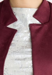 Burgunderfarbener Blazer mit elegantem Kragen, kombiniert mit einem hellsilbernen, strukturierten Top mit subtilen Mustern. Weicher Stoff mit glatter Oberfläche.