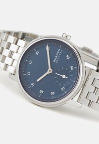 Stříbrné nerezové hodinky s kulatým námořnickým modrým ciferníkem. Obsahují minimalistické hodiny a minutové ručičky, malý subciferník a spojený náramek.