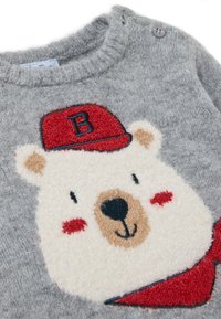 Pull en tricot gris avec un design de ours texturé. L'ours porte une casquette rouge et a une écharpe. Deux boutons-pression à l'épaule.