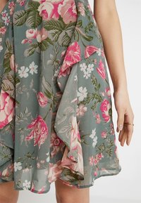 Zielona sukienka w kwiatowy wzór z różowymi, białymi i jasnoróżowymi kwiatami, z warstwowymi falbanami przy hemie, pokazana na dolnej części ciała osoby.