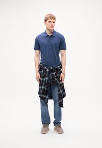 Joven de pie que lleva una camiseta polo azul, jeans, zapatillas beige y una camisa de cuadros en negro y azul atada a la cintura.