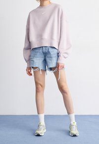 Osoba w jasnoróżowej bluzie Nike, podartych jeansowych szortach, białych skarpetkach i białych butach sportowych Nike stoi na niebieskiej podłodze.