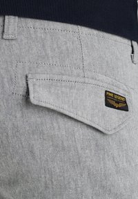 Tejido de mezclilla gris con una superficie texturada. El bolsillo trasero en ángulo presenta una etiqueta negra con el texto "PME LEGEND" y un gráfico de alas.