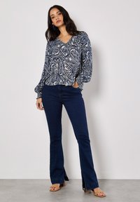 Bluse mit marineblauem und cremefarbenem Paisleymuster, V-Ausschnitt, langen Puffärmeln und gerüschtem Saum, kombiniert mit dunkelblauen Schlagjeans und hellbraunen Absatzsandalen.