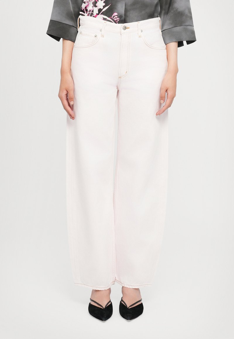 Φαρδιά τζιν σε ανοιχτό ροζ χρώμα με διπλές ραφές, εμπρόσθιες τσέπες και κουμπί στη ζώνη μέσης, συνδυασμένα με μαύρες γόβες μυτερής μύτης.