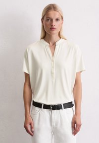 Polo shirt crème à manches courtes avec un col chinois, comportant une patte de boutonnage. Porté avec un jean blanc et une ceinture noire. Texture lisse.