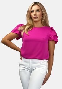 Jasnoróżowa bluzka z krótkimi falbaniastymi rękawami, okrągłym dekoltem i gładką teksturą, w połączeniu z białymi dopasowanymi spodniami.