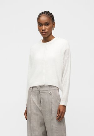 Γυναίκα με πλεξούδες, φορεί μια λευκή ζακέτα με κουμπιά και ψηλόμεσο μπεζ καρό παντελόνι, στέκεται μπροστά σε απλό ανοιχτόχρωμο φόντο.