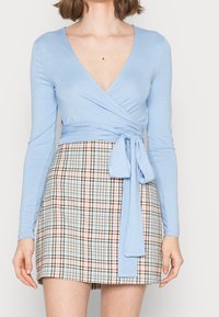 Hellblaues Langarm-Wickeloberteil mit tiefem V-Ausschnitt und Bindungsdetail. Kombiniert mit einem karierten Minirock in Beige-, Braun- und Blautönen.