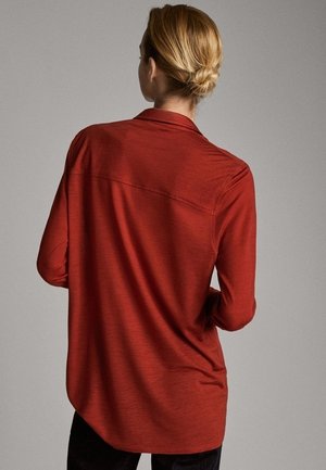 Femme aux cheveux blonds attachés en chignon, portant une chemise ample à manches longues couleur rouille, vue de dos sur un fond neutre.
