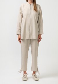 Světlý béžový outfit obsahující bundu na zip s stojáčkem a odpovídající kalhoty se šňůrkou. Měkká látka s hladkou texturou.