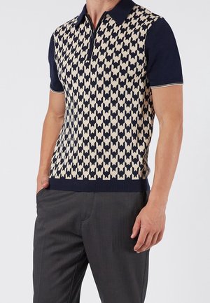 Mann trägt dunkelblaues Polo-Shirt mit kurzen Ärmeln mit beigem und dunkelblauem geometrischem Muster und dunkelgrauer Hose, steht mit einer Hand in der Tasche.