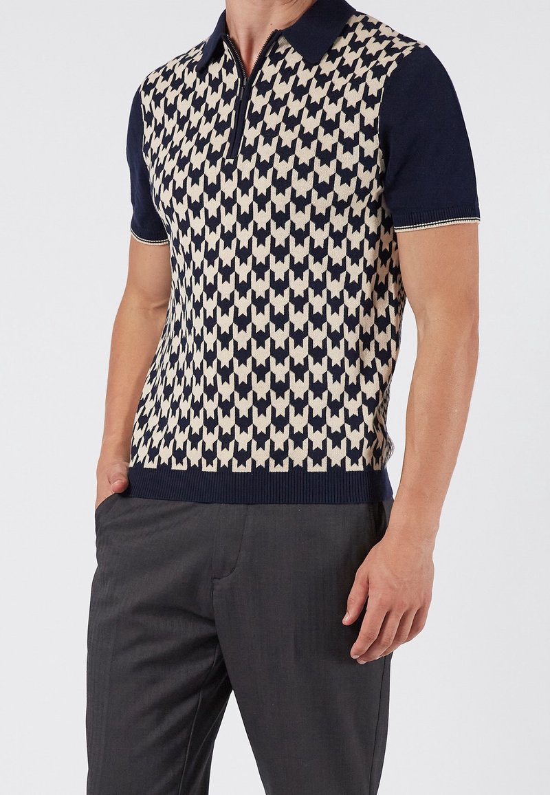 Homme portant un polo manches courtes bleu marine avec un motif géométrique beige et bleu marine et un pantalon gris foncé, debout avec une main dans la poche.