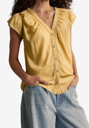 Kobieta ubrana w żółtą, krótki rękaw, zapinaną na guziki bluzkę z marszczonymi ramionami oraz jasnoniebieskie, wysokie spodnie jeansowe, z rękami w kieszeniach.