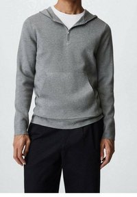 Un sweat à capuche gris zippé avec une poche avant, fabriqué en tissu doux, comporte une capuche, des manches longues et une coupe décontractée. Associé à un pantalon noir.