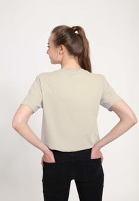 Krátký béžový tričko s kulatým výstřihem, vyrobené z měkké bavlny, v kombinaci s černými džíny; model stojí s rukama v kapsách.