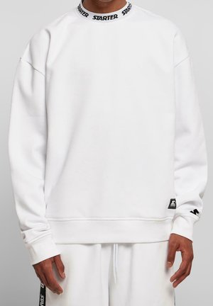 Mężczyzna w białej bluzie z napisem "STARTER" na kołnierzu, małą naszywką z logo przy dole oraz w białych spodniach z bocznym paskiem.