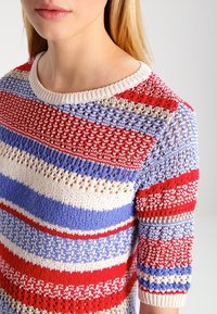 Neulepaita, jossa on vaakasuoria raitoja punaisessa, sinisessä ja kermanvärisessä. Ominaisuuksiin kuuluu pyöreä kaula-aukko ja lyhyet hihat, joissa on teksturoitu kuvio.