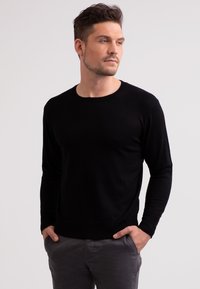 Schwarzer Strickpullover mit langen Ärmeln, runder Ausschnitt und geripptem Saum. Glatte Textur und figurnaher Stil, getragen mit grauen Hosen.