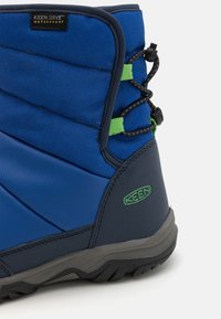 Blauer wasserfester Stiefel mit strukturiertem Finish, ausgestattet mit schwarzen und grünen Schnürsenkeln. Gummisohle und gepolsterter Kragen für zusätzlichen Halt. Logo auf der Seite sichtbar.