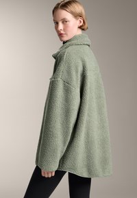 Overgrote groene fleece jas met een textuurfinish, voorzien van een kraag en verlaagde schouders, gecombineerd met zwarte fitted leggings.