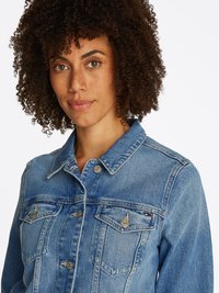 Jaqueta de denim azul com gola, fecho frontal de botões, dois bolsos no peito com aba e detalhes metálicos. Corte e textura clássicos.