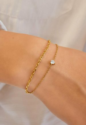 Poignet portant deux bracelets délicats en or, dont un avec une petite pierre précieuse claire en forme de cœur, sur un fond de tissu clair.