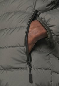 Gray padded jacket featuring a zippered pocket and elastic cuffs. The fabric has a smooth texture with horizontal stitching across the surface.