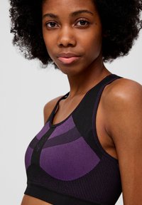 Lila und schwarze Sport-BH mit einem strukturierten Muster, breiten Trägern und einem engen Sitz. Der Stoff scheint dehnbar und hat eine glatte Oberfläche.