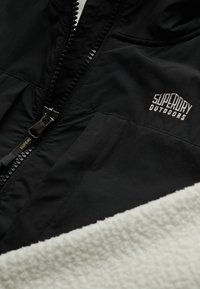 Veste noire Superdry Outdoors avec fermeture éclair partiellement fermée, avec une doublure en polaire blanche au bas.