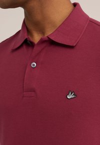 Burgundské polo tričko vyrobené z texturované látky, s dvouknoflíkovou légou a malým černým vyšitým logem ptáka na hrudi.