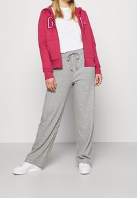 Femme portant un sweat à capuche rouge zippé, un t-shirt blanc, un pantalon de jogging gris avec cordon de serrage, et des baskets blanches, debout devant un fond uni.