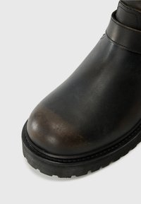 Bota de cuero negro con punta redondeada, suela gruesa tipo lug y detalle de tira cosida cerca del tobillo.