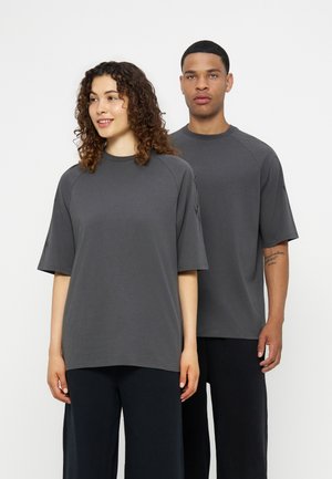 Due adulti con magliette larghe grigio scuro e pantaloni neri stanno in piedi su uno sfondo bianco neutro.