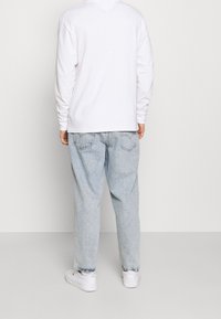 Person, der ein weißes langärmliges Shirt, hellblaue Jeans und weiße Sneaker trägt, steht vor einem einfarbigen hellen Hintergrund.