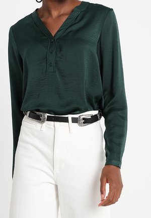 Femme portant un chemisier vert foncé à manches longues rentré dans un pantalon blanc taille haute avec une ceinture noire avec des boucles argentées.