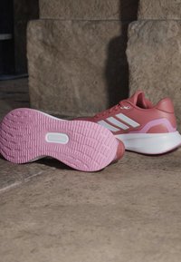 Paire de chaussures de sport roses avec des rayures blanches et des semelles intermédiaires blanches épaisses, placées sur un sol en pierre contre un mur en pierre.